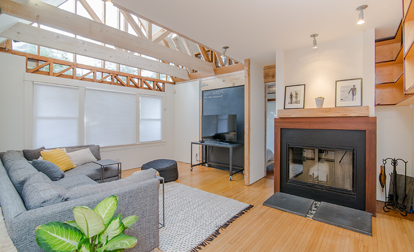 A modern living room with a cozy gray couch, a fireplace, and large windows showcasing wooden beams.